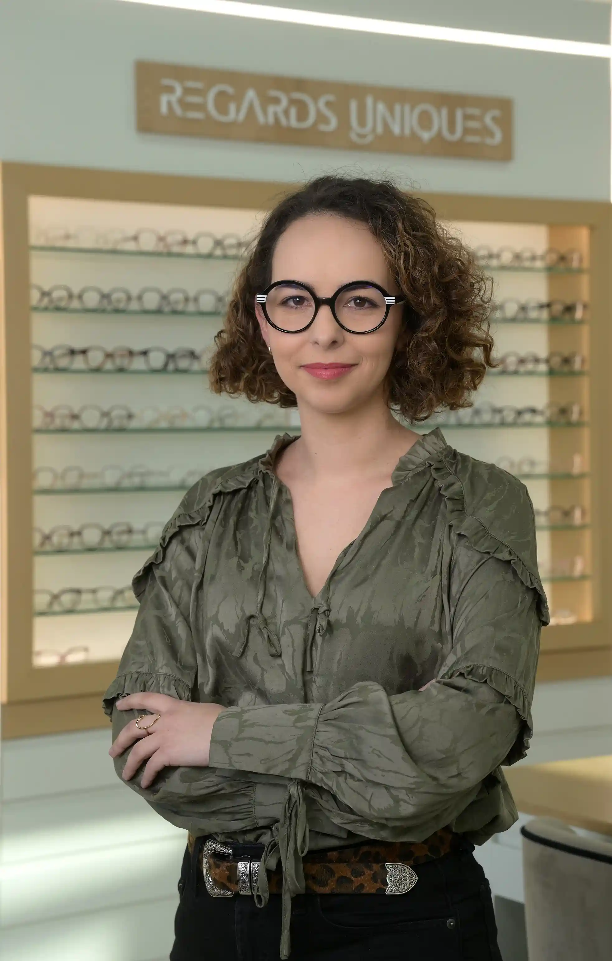 Portrait d’une opticienne professionnelle dans la boutique Regards Uniques, posant avec assurance devant un mur de montures de lunettes