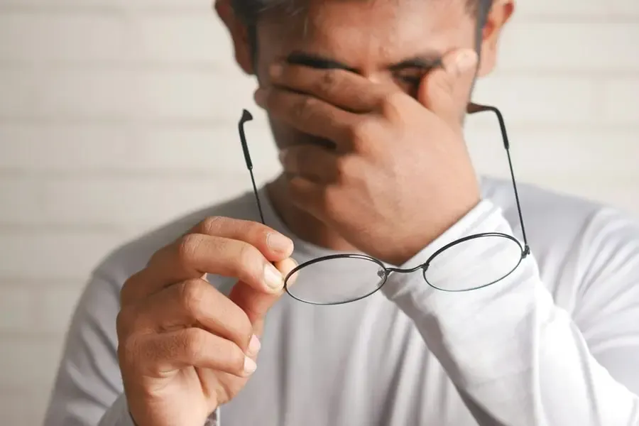 Homme se frottant les yeux en tenant ses lunettes, illustrant la fatigue visuelle et le besoin d’une solution optique adaptée chez Regards Uniques