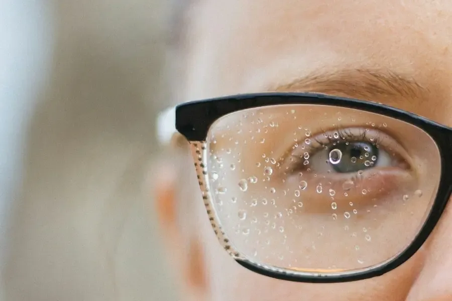 Gros plan sur un œil derrière des lunettes avec des gouttelettes d’eau, illustrant les défis de la vision par temps humide et les solutions optiques proposées par Regards Uniques