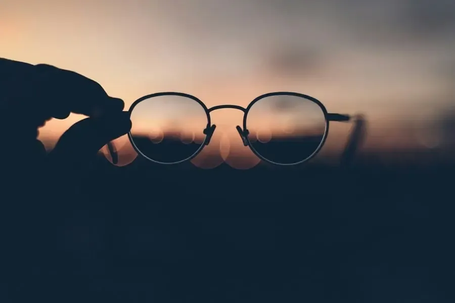 Paire de lunettes fines tenue face à un coucher de soleil, symbolisant la clarté visuelle et la beauté du regard selon Regards Uniques