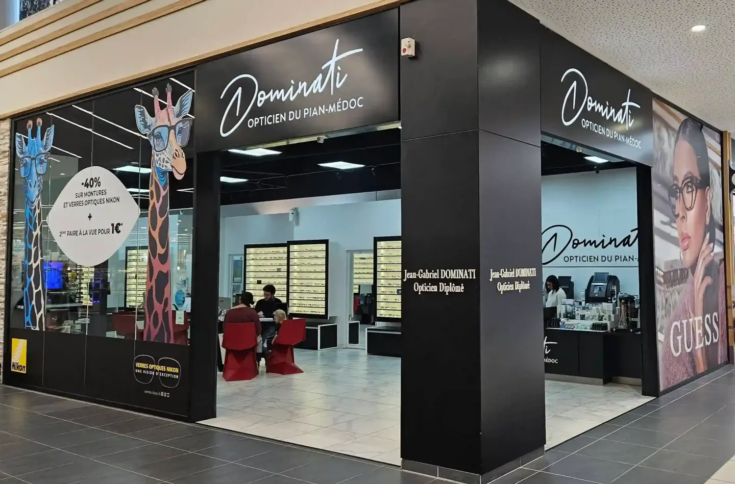 Vue extérieure d'une boutique d'optique 'Dominati Opticien du Pian-Médoc' dans un centre commercial. Le magasin est bien éclairé et affiche une promotion sur les lunettes, avec des présentoirs de lunettes visibles à l'intérieur. Une grande affiche de la marque Guess est également présente.
